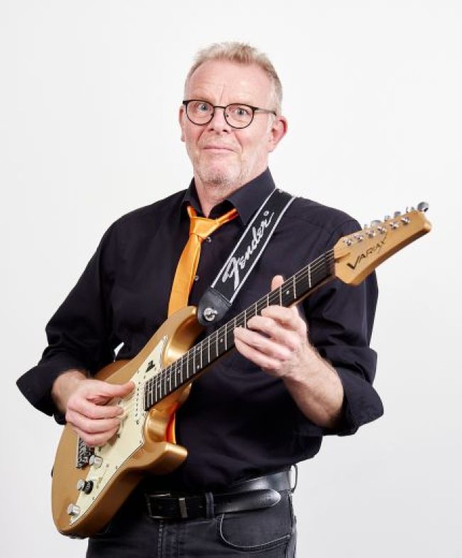 Ein Mann mit Brille und schwarzem Shirt hält eine E-Gitarre in der Hand und lächelt begeistert.