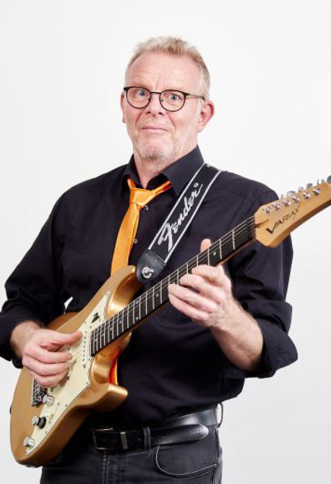 Ein Mann mit Brille und schwarzem Shirt hält eine E-Gitarre in der Hand und lächelt begeistert.