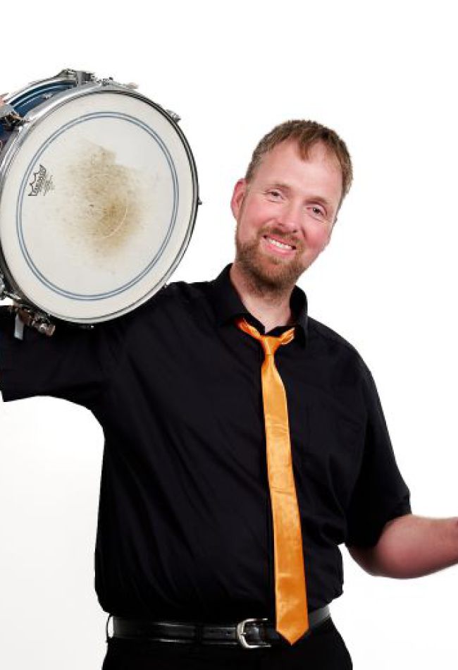 Ein Mann in schwarzem Hemd und Krawatte hält ein Schlagzeug und Sticks in der Hand, bereit zum Spielen.