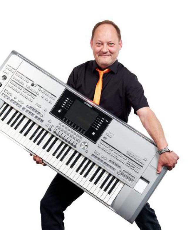 Ein Mann steht mit einem großen elektronischen Keyboard in der Hand und lächelt in die Kamera.