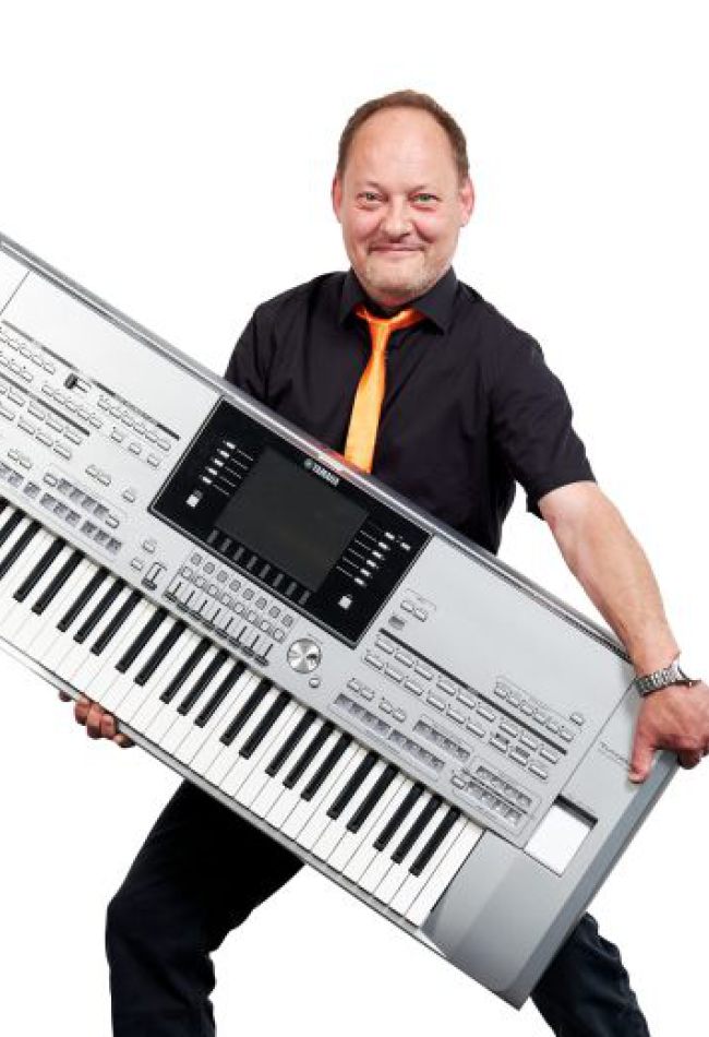 Ein Mann steht mit einem großen elektronischen Keyboard in der Hand und lächelt in die Kamera.