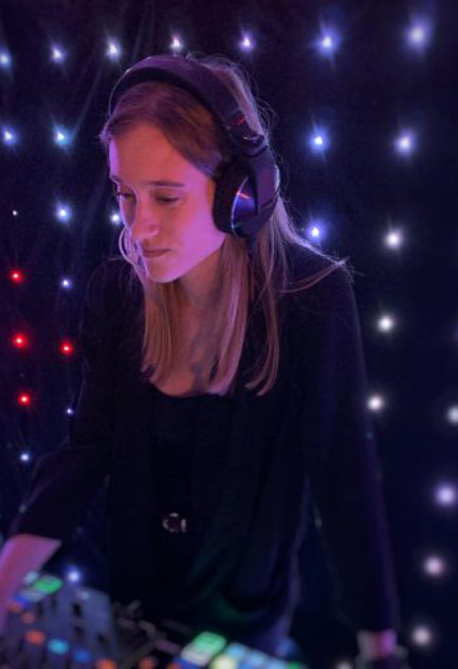 Eine DJane mit Kopfhörern und einem schwarzem Outfit steht an einem Mischpult.